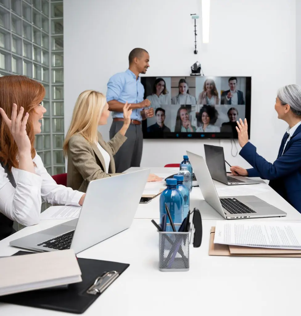 Barrister Suites Office Lease Four people in a conference room with laptops wave at a screen showing a video call with multiple participants. One man stands near the screen, while three women sit at the table, engaging in the meeting.