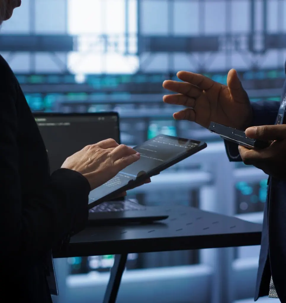 Barrister Suites Office Lease Two people in business attire discuss information displayed on a tablet and smartphone, standing in a modern server room with computer monitors and server racks in the background.