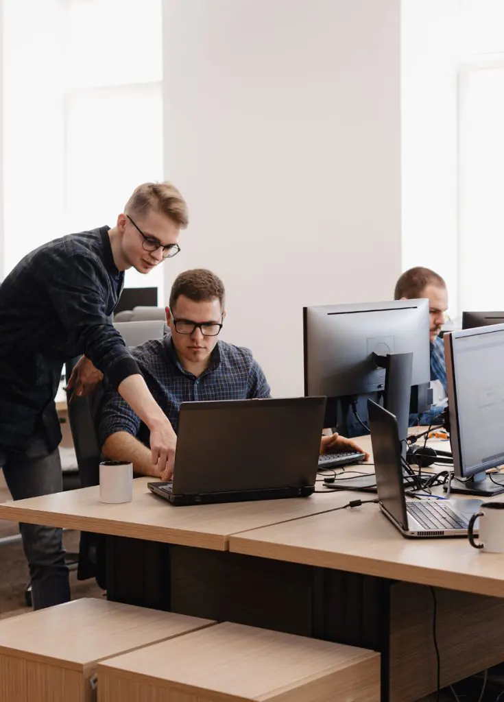 Barrister Suites Office Lease Two men at an office workstation focus on a laptop; one stands and points at the screen while the other sits. Other employees work at nearby desks with computers and coffee mugs visible.