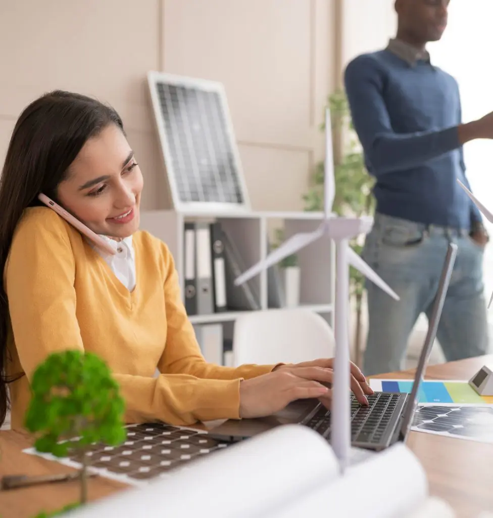 Barrister Suites Office Lease A woman in a yellow sweater talks on the phone and types on a laptop at a desk with a wind turbine model and a small tree model. A man stands in the background near blueprints and a solar panel.
