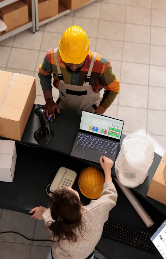 Barrister Suites Office Lease A person in a hard hat and work overalls stands at a counter with boxes, while another person sits at a desk with a laptop, phone, and a yellow hard hat, discussing or reviewing information.
