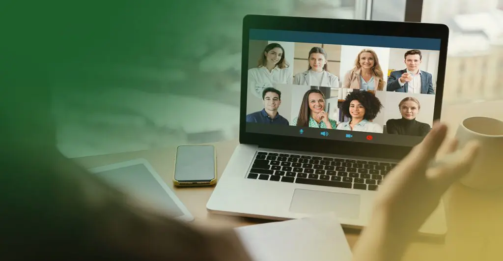 Barrister Suites Office Lease A person sits at a desk with a laptop open, participating in a video call with eight people. A smartphone, tablet, and coffee cup are also on the desk. The persons hand is raised toward the screen.
