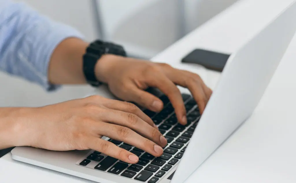 Barrister Suites Office Lease Close-up of a persons hands typing on a laptop keyboard, with a smartphone placed on the white desk beside them. The person is wearing a dark wristwatch and a light blue long-sleeve shirt.