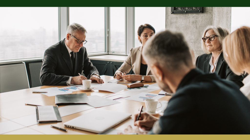 Barrister Suites Office Lease Five business professionals sit around a conference table with documents, charts, and laptops, engaged in a meeting in a modern office with large windows.