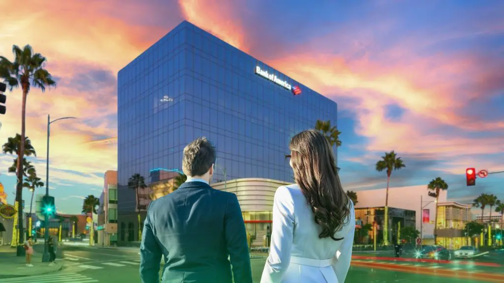 Barrister Suites Office Lease A man and a woman, seen from behind, stand at an intersection facing a glass Bank of America building, with palm trees and a colorful sunset sky in the background.