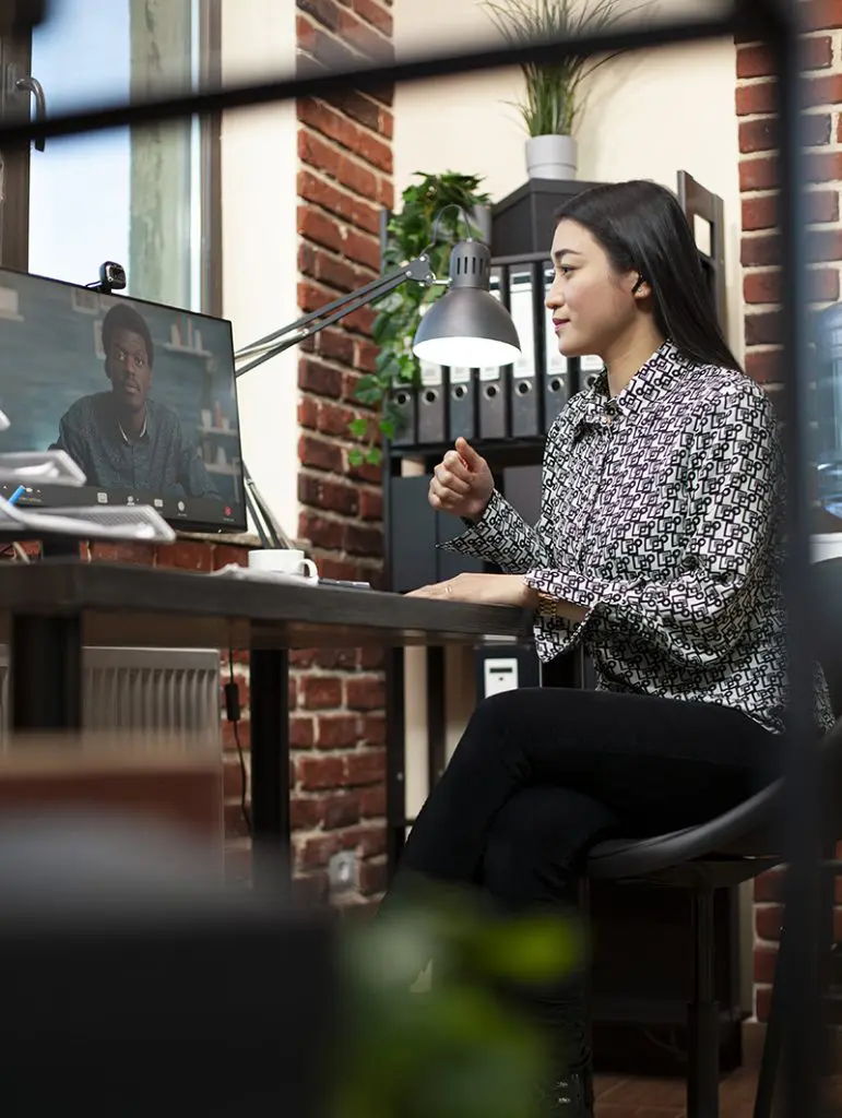 Barrister Suites Office Lease A woman sits at a desk in a modern office, participating in a video call with a man shown on her computer monitor. The workspace has exposed brick walls, shelves, and plants.
