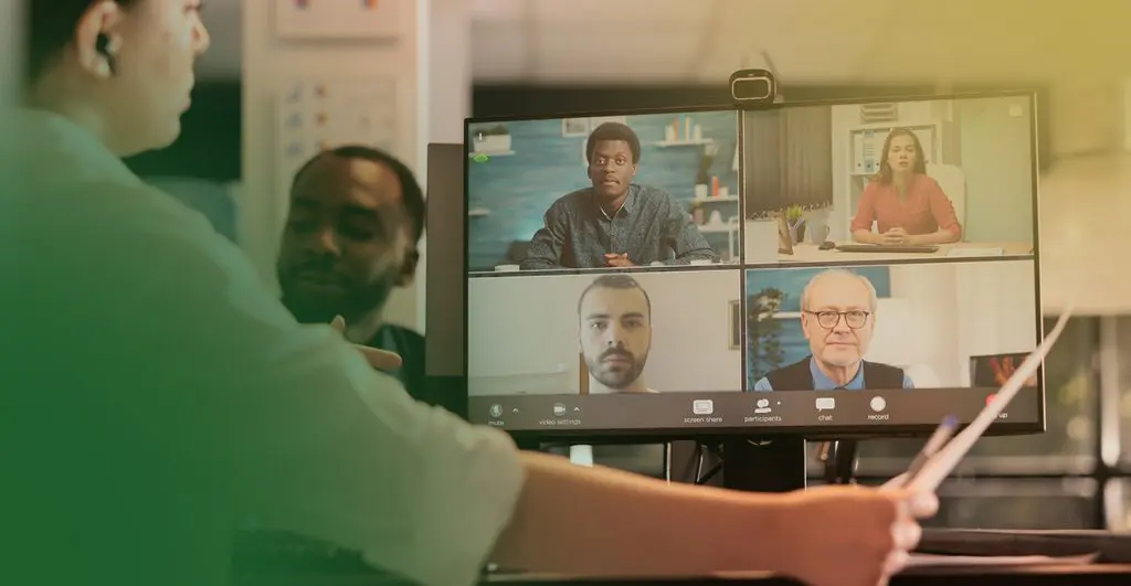 Barrister Suites Office Lease Two people in an office talk in front of a computer monitor showing a video call with four participants, each in a separate window, engaged in a virtual meeting.