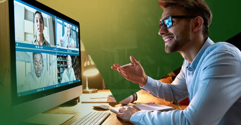 Barrister Suites Office Lease A smiling man wearing glasses sits at a desk with a pen in hand, participating in a video conference call with four people shown on his computer screen. The desk is lit by a lamp and has a keyboard and mouse.