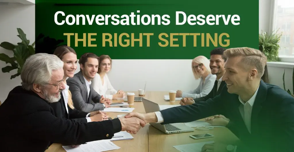 Barrister Suites Office Lease A group of businesspeople sit around a conference table, smiling as two men in the foreground shake hands. The text above reads, “Conversations Deserve THE RIGHT SETTING.”.