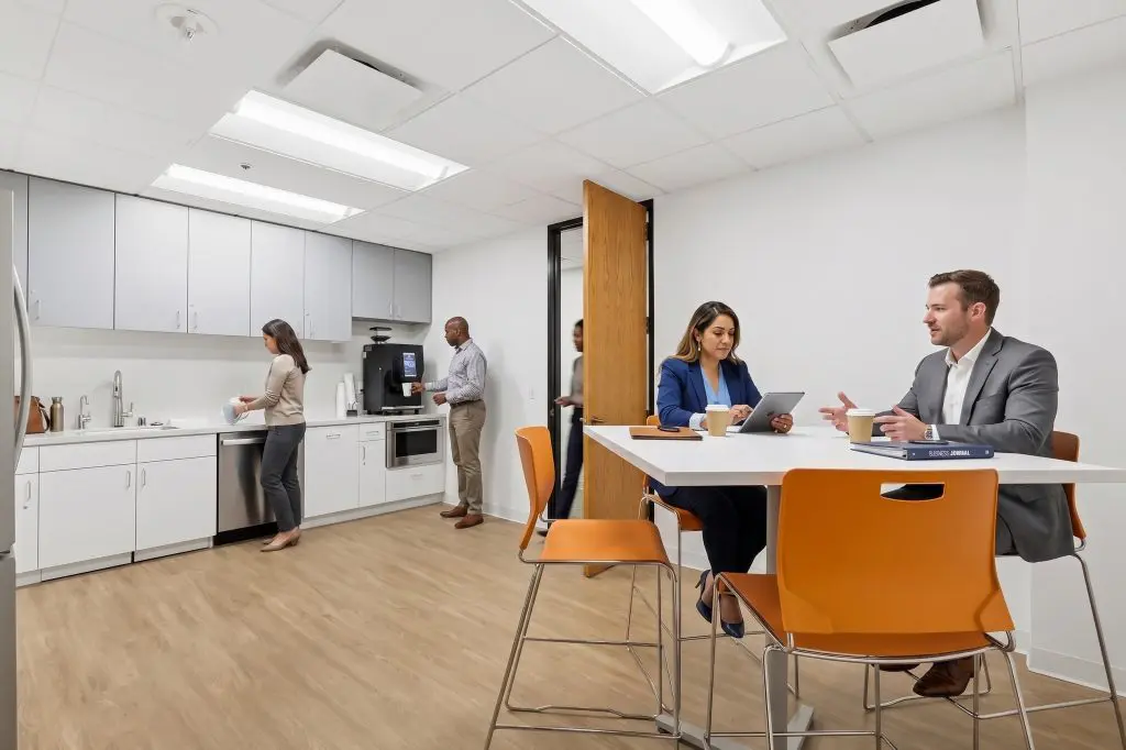 Barrister Suites Office Lease Four people are in a modern office kitchen; two are seated at a high table talking, while two others prepare drinks at the counter. The space has white cabinets, appliances, and orange chairs.