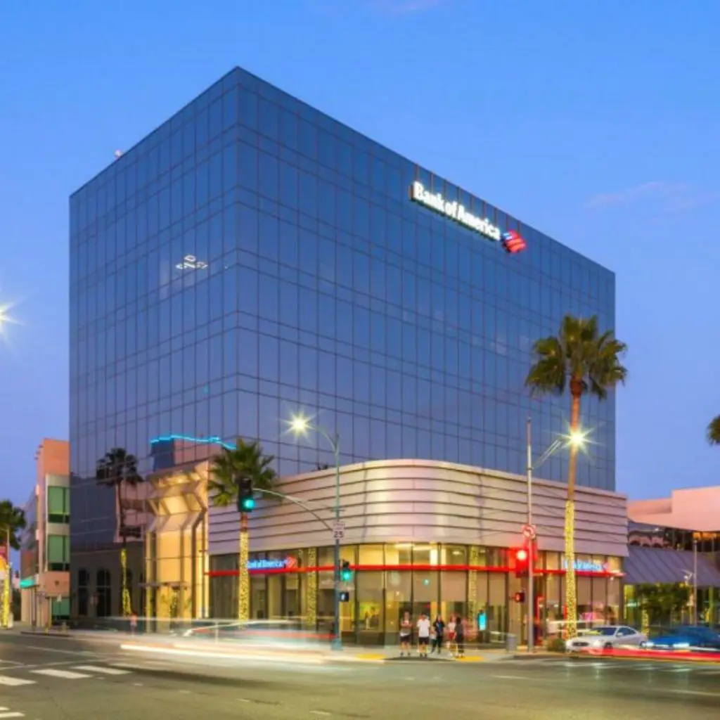 Barrister Suites Office Lease A modern glass office building with a Bank of America branch on the ground floor, illuminated at dusk, located at a busy street intersection with palm trees and light trails from passing cars.