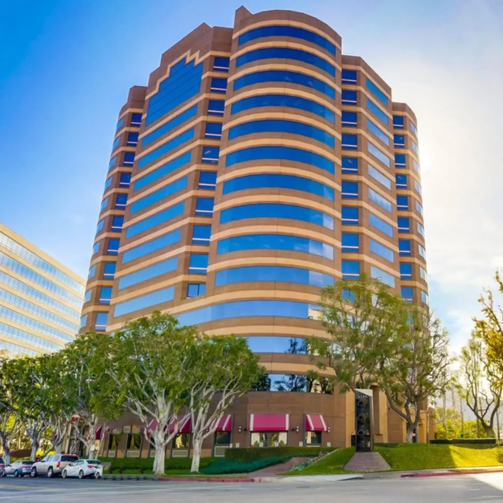 Barrister Suites Office Lease A tall, modern office building with curved edges and alternating bands of tan and blue glass, surrounded by green trees and parked cars, under a clear sky.