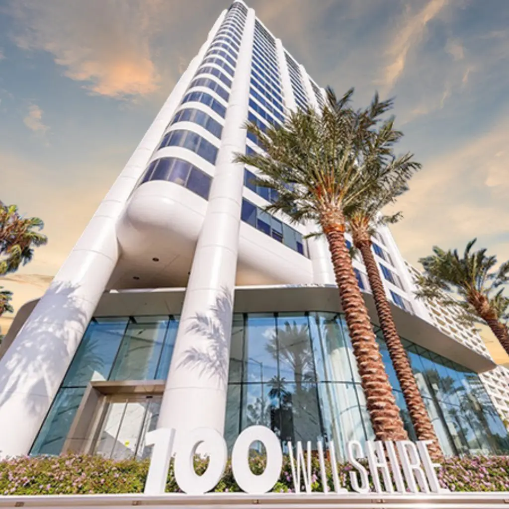 Barrister Suites Office Lease A tall, modern glass and white building with palm trees in front. Large white numbers and letters at the entrance read 100 WILSHIRE. The sky is partly cloudy with a warm, golden sunset.