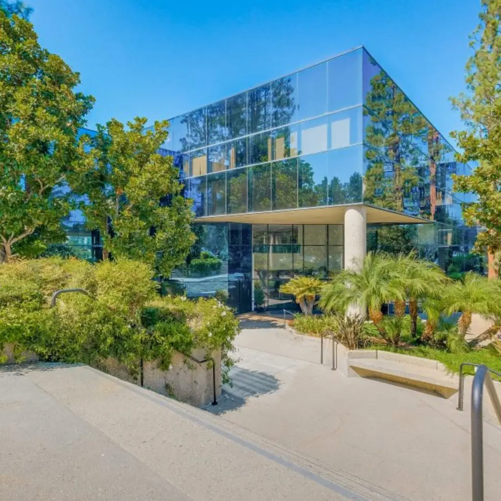 Barrister Suites Office Lease A modern glass office building surrounded by lush green trees and landscaping, with a concrete stairway leading to the entrance under a clear, blue sky.