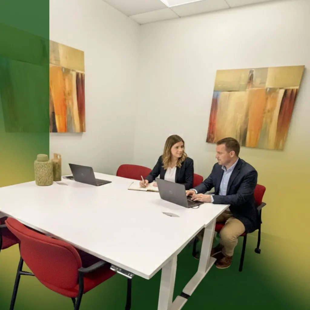 Barrister Suites Office Lease Two people in business attire sit at a white table in a modern office, working on laptops and taking notes. Abstract paintings decorate the walls, and red chairs surround the table.