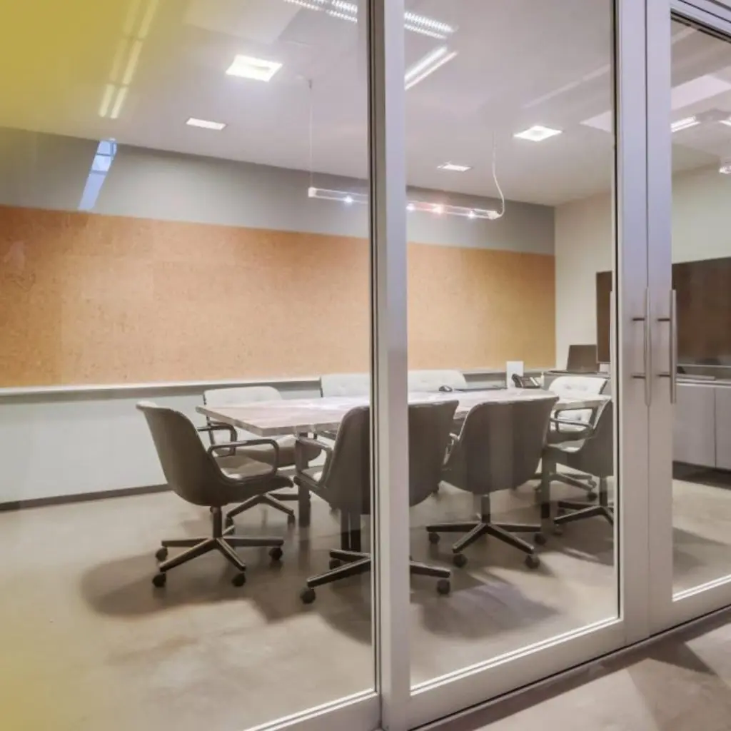 Barrister Suites Office Lease A modern conference room with glass doors, a rectangular table, several office chairs on wheels, and a large cork board on the back wall. The room is well-lit and has a minimalist design.