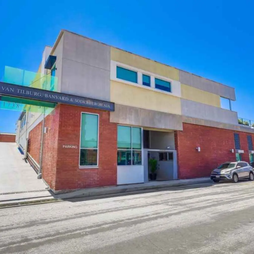 Barrister Suites Office Lease Modern two-story building with a mix of red brick and concrete, large windows, and a car parked outside. A sign with business names is mounted on the upper corner. The sky is clear and blue.