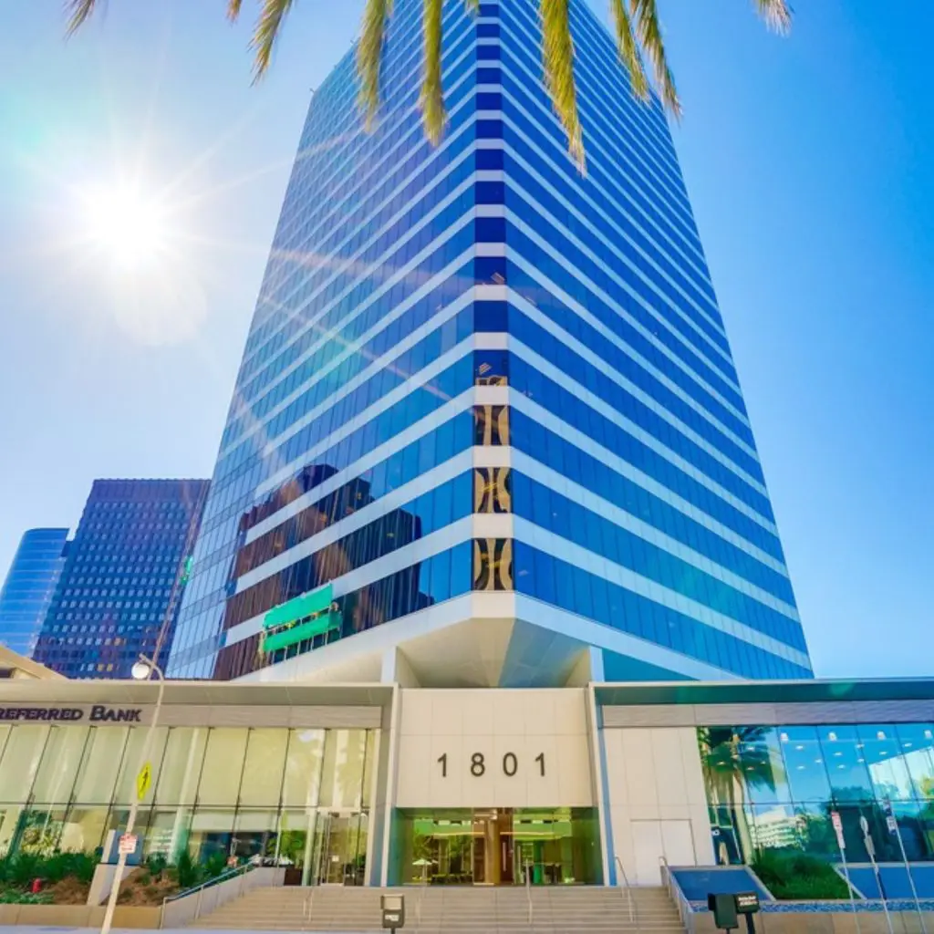 Barrister Suites Office Lease A tall, modern glass office building with the number 1801 above the entrance, bathed in sunlight with palm tree leaves at the top and a clear blue sky in the background.