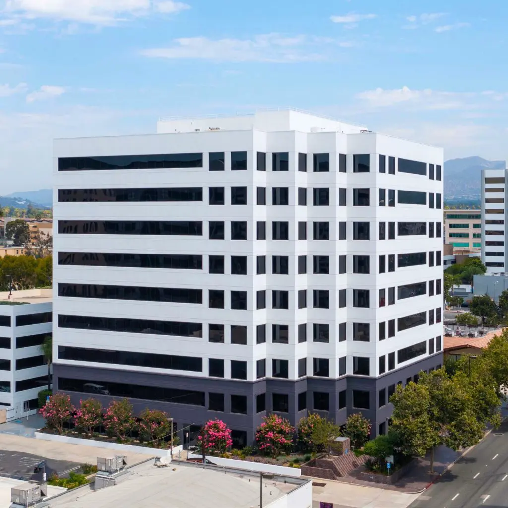Barrister Suites Office Lease A modern, white and black office building with multiple floors, surrounded by trees and smaller buildings, under a partly cloudy sky.