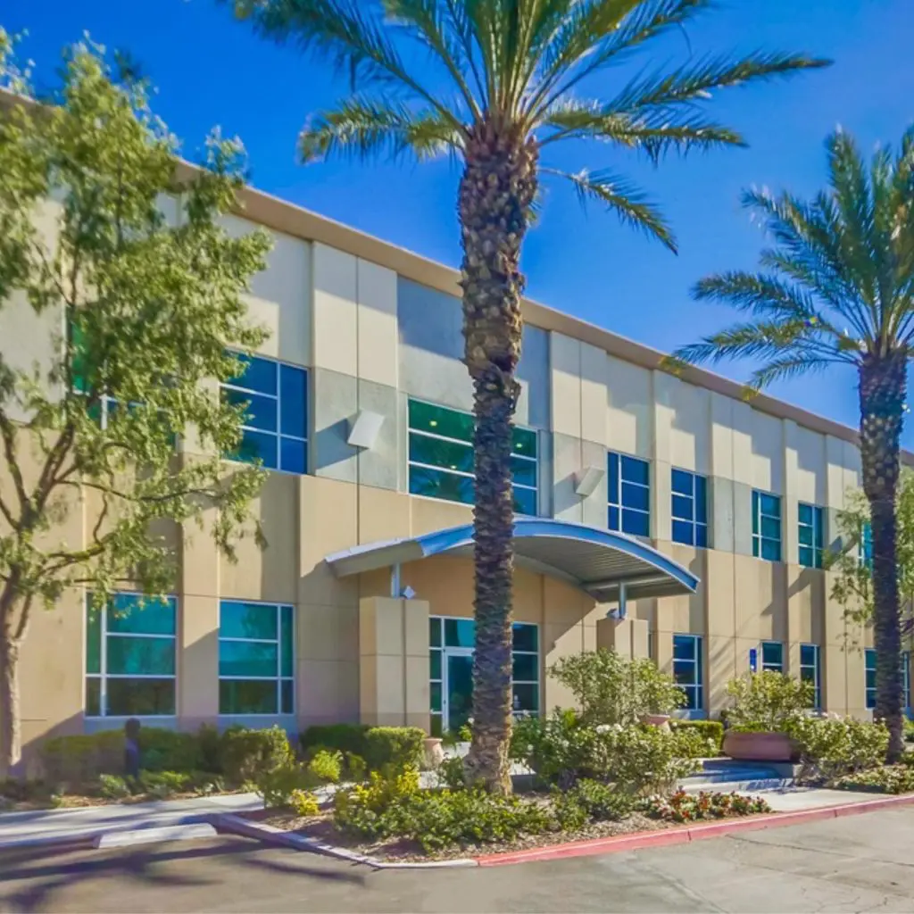 Barrister Suites Office Lease A modern two-story building with large windows, surrounded by palm trees and landscaped greenery, under a clear blue sky. The entrance features a curved metal awning.