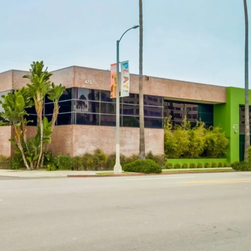 Barrister Suites Office Lease A modern brick office building with green accents and large tinted windows sits on a street corner. Tall palm trees and lush landscaping surround the building, and a streetlamp with banners stands in front.