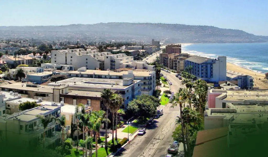 Barrister Suites Office Lease A coastal cityscape with palm trees, mid-rise buildings, and a road running parallel to a sandy beach and the ocean; hills are visible in the hazy background.
