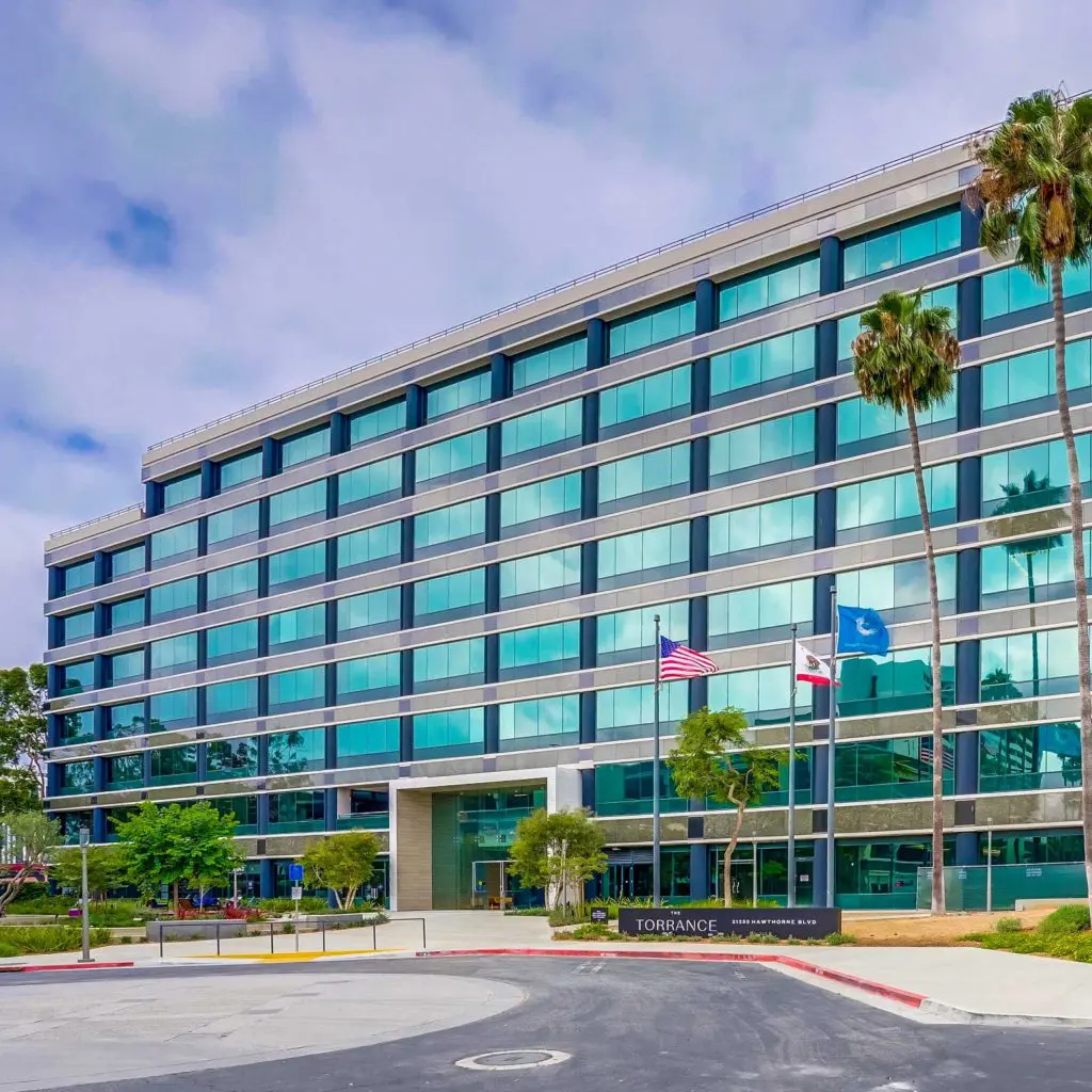 Barrister Suites Office Lease A modern glass office building with six stories, featuring rows of windows, a main entrance, and three flagpoles with the U.S., California, and city flags in front. Palm trees and greenery line the street outside.