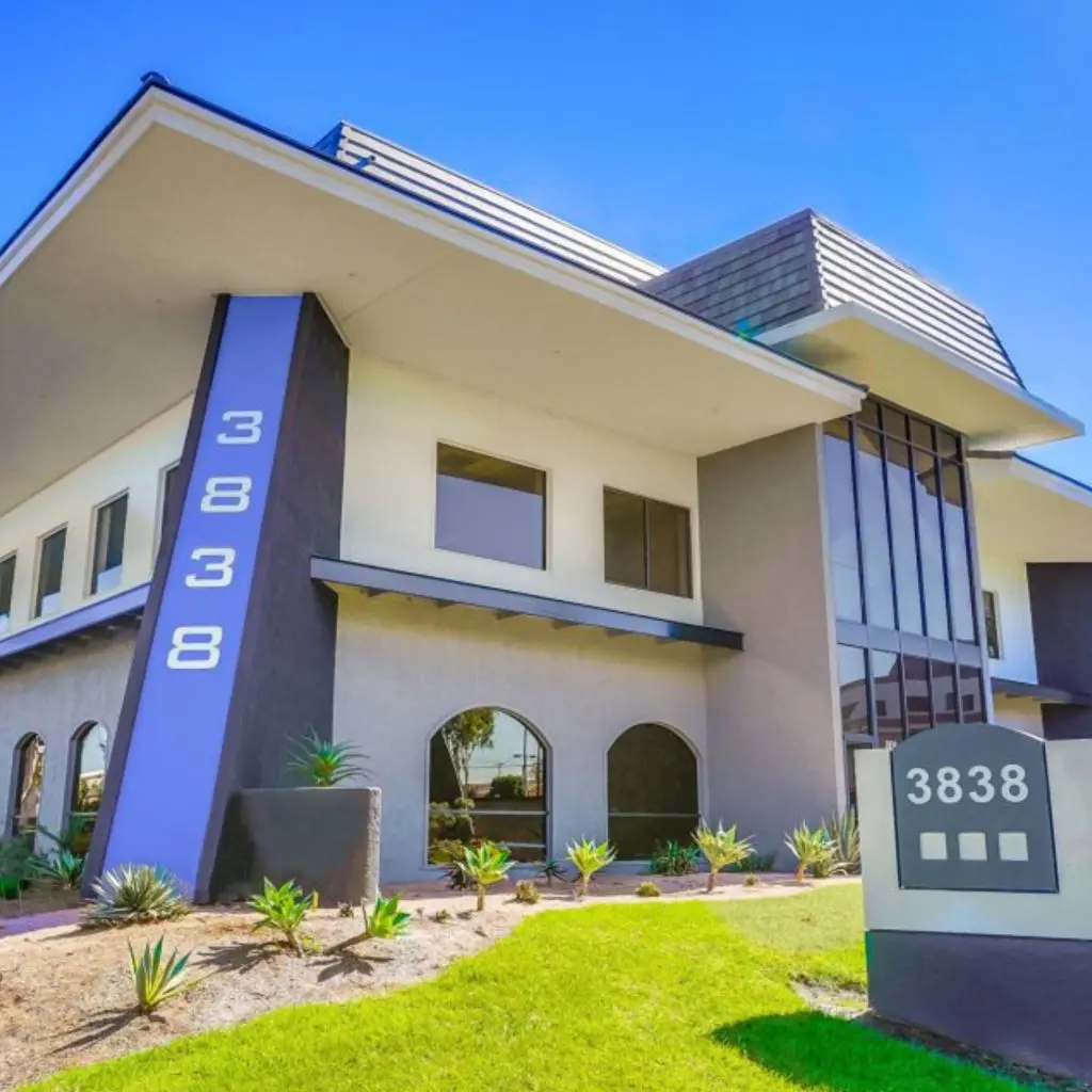 Barrister Suites Office Lease Modern two-story commercial building with large windows, grey and white exterior, and the number 3838 displayed vertically on the facade and on a sign in front. Desert landscaping with small plants is visible.
