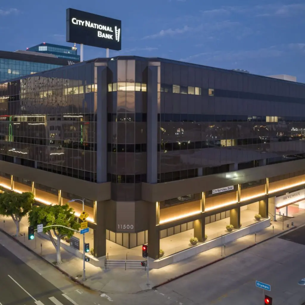 Barrister Suites Office Lease A modern multi-story office building at dusk with a prominent City National Bank sign on the roof and illuminated street corners at the intersection of Olympic Blvd and a cross street. The address 11500 is visible on the building.