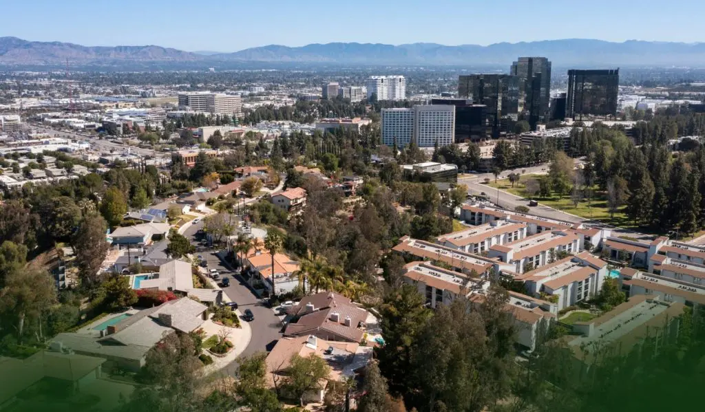 An aerial view of a suburban neighborhood with winding streets and trees, beyond which there are office towers and distant mountains under a clear blue sky.