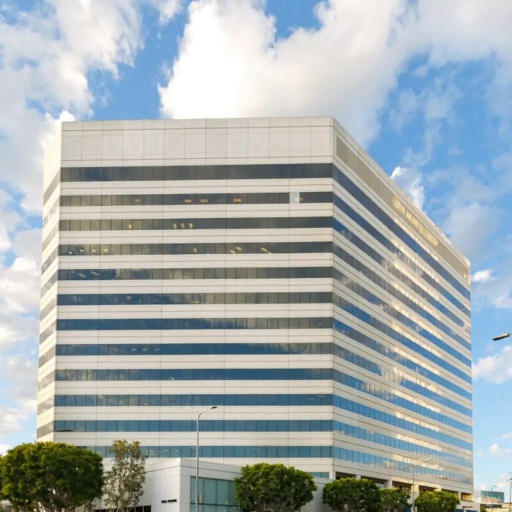 Barrister Suites Office Lease A tall, modern office building with many horizontal windows and white trim stands against a partly cloudy sky, surrounded by small green trees and bushes at its base.
