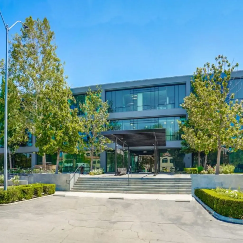 Barrister Suites Office Lease A modern glass office building with three stories, surrounded by trees and landscaping, and a staircase leading up to the main entrance from an empty parking lot.