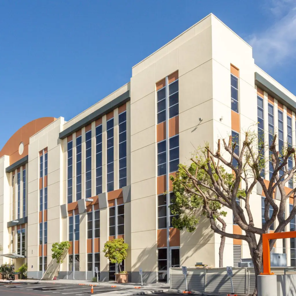 Barrister Suites Office Lease A modern, multi-story office building with tall windows and orange accents, surrounded by a few small trees, under a clear blue sky.