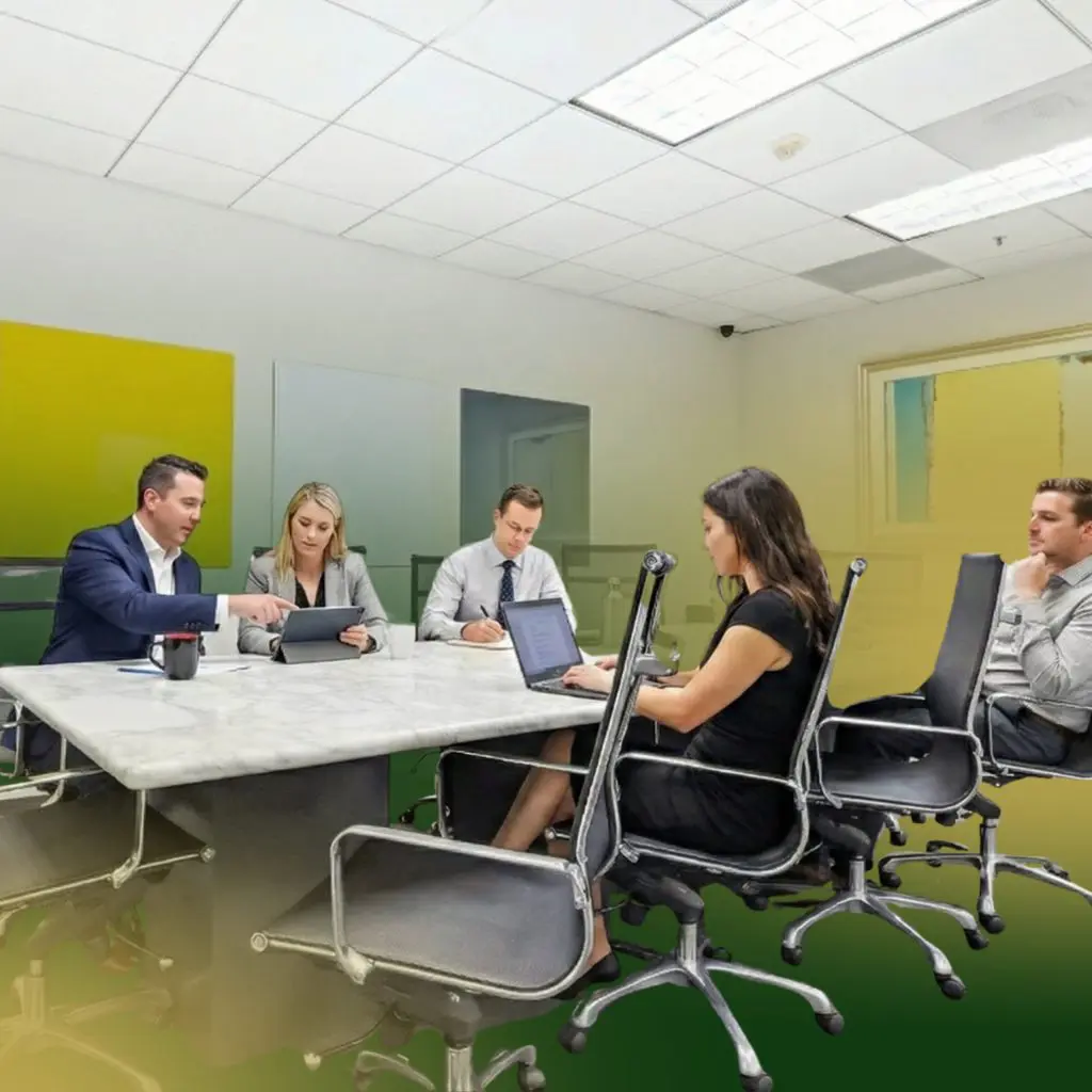 Barrister Suites Office Lease Five people in business attire are seated around a conference table in a modern office, engaged in a meeting. Two work on laptops, while others discuss and review documents. The room has abstract wall art and overhead lighting.