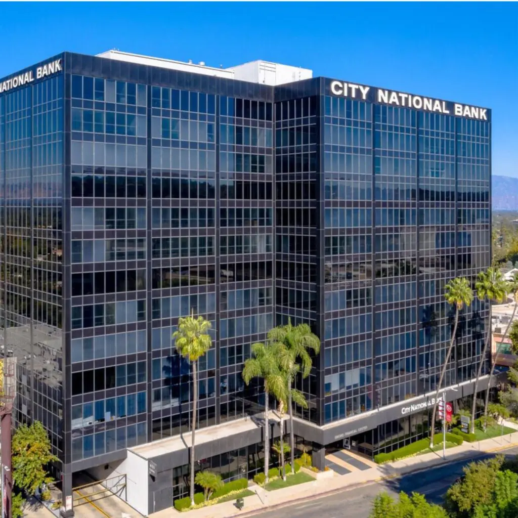 Barrister Suites Office Lease A modern, multi-story glass office building with City National Bank signage on the top, surrounded by palm trees and reflecting the blue sky.