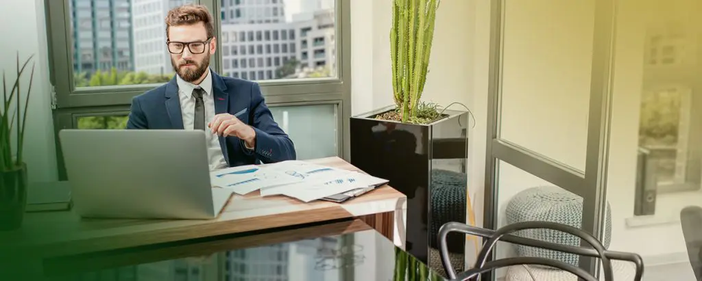 Barrister Suites Office Lease A man in a suit and glasses sits at a desk in a modern office, working on a laptop with documents and charts spread out. A large window and a tall potted cactus are in the background.