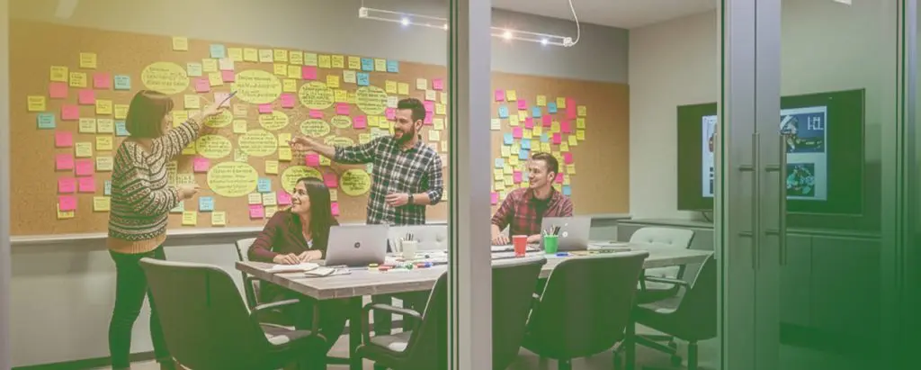 Barrister Suites Office Lease Four people collaborate in a conference room with laptops and notebooks; colorful sticky notes cover a large board on the wall behind them. Two people are standing and pointing at the notes, while two are seated and smiling.
