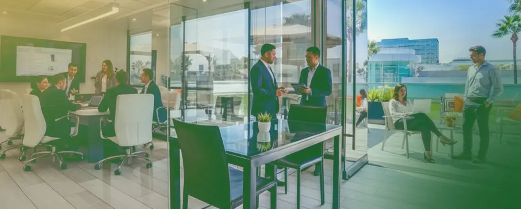 Barrister Suites Office Lease A modern office with people meeting indoors around a table and others talking or relaxing outside on a sunny terrace with palm trees and glass buildings in the background.
