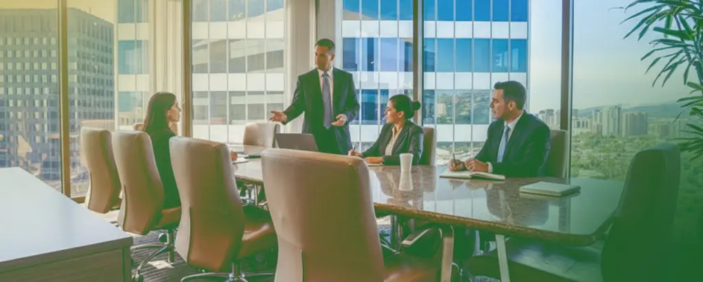 Barrister Suites Office Lease Four people in business attire sit around a conference table in a modern office with large windows, city buildings visible outside. One man stands and speaks to the group while the others listen attentively.
