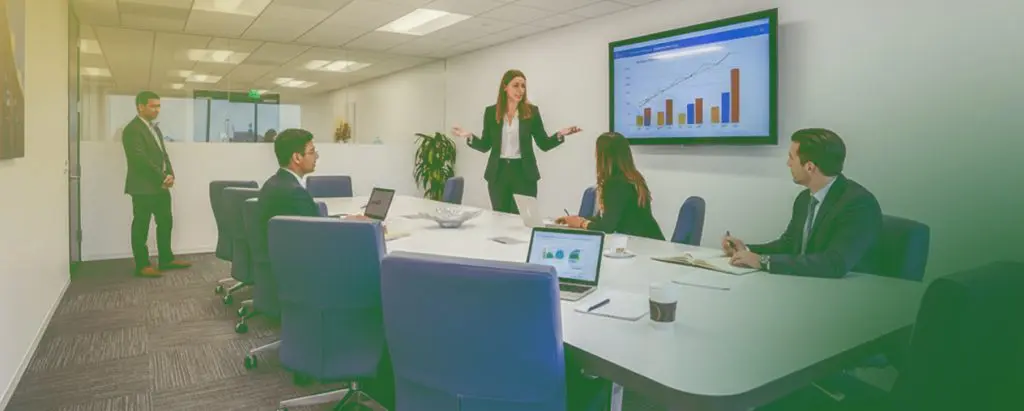 Barrister Suites Office Lease Five people in a modern conference room, with one person presenting a bar and line graph on a screen while others listen and use laptops.