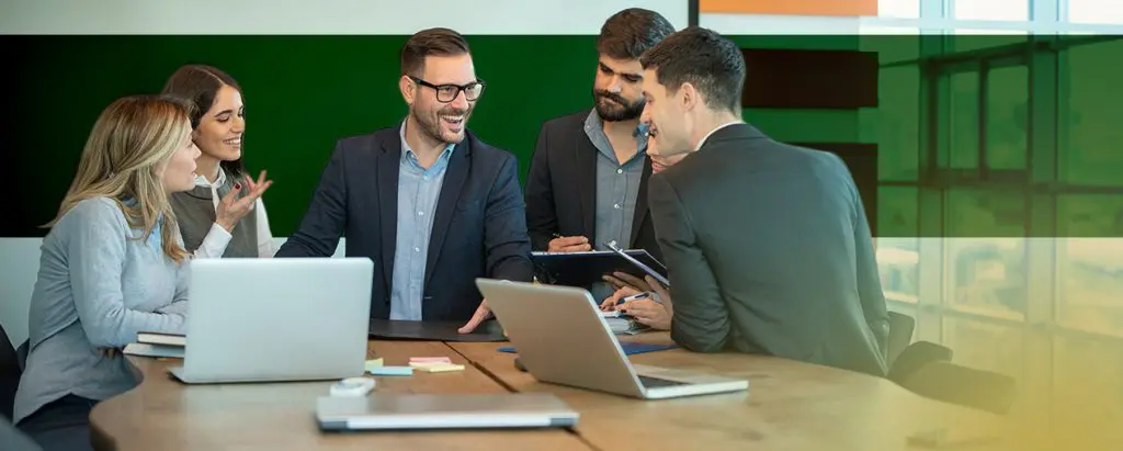 Barrister Suites Office Lease Five people in business attire sit around a conference table with laptops, notebooks, and papers, engaged in a lively discussion and smiling in a modern office setting.