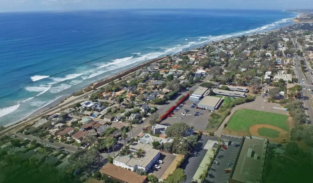 Barrister Suites Office Lease Aerial view of a coastal town with houses, a baseball field, roads, and a long sandy beach with waves; the ocean stretches along the left side of the image, while the town extends along the shoreline to the right.