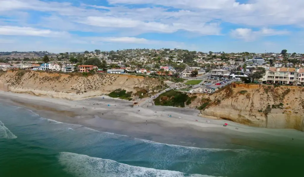 Barrister Suites Office Lease Aerial view of a beach with gentle waves, people scattered on the sand, and steep cliffs topped with houses, buildings, and greenery beneath a partly cloudy sky.