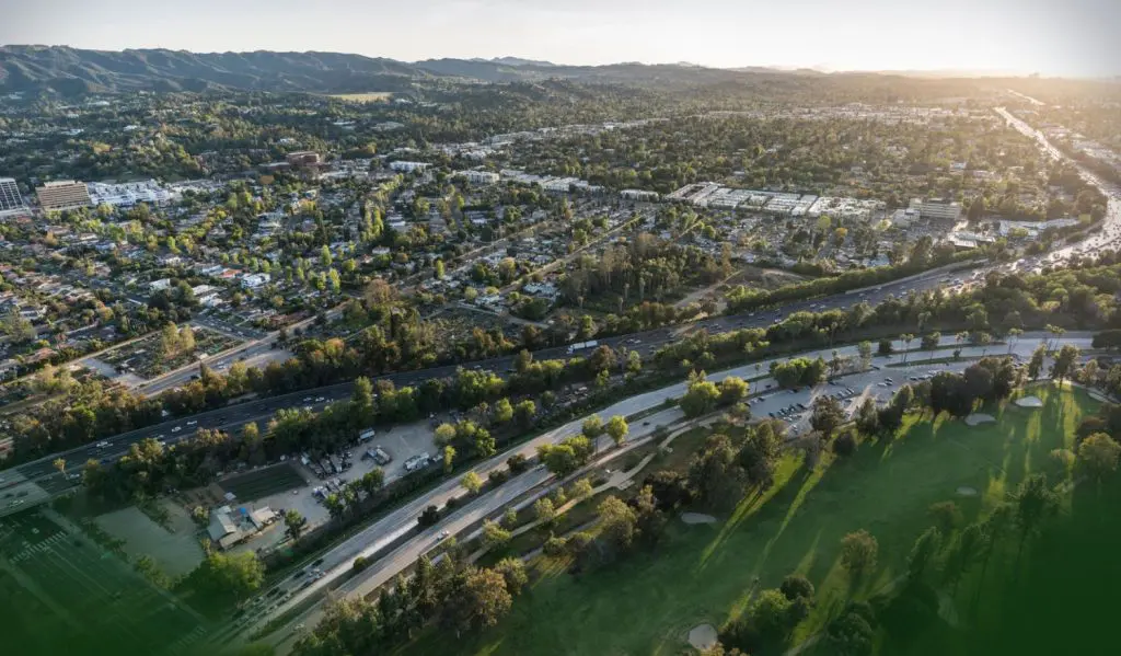 Barrister Suites Office Lease Aerial view of a suburban neighborhood with tree-lined streets, a highway, parking lots, and green park space in the foreground, set against distant hills under a setting sun.