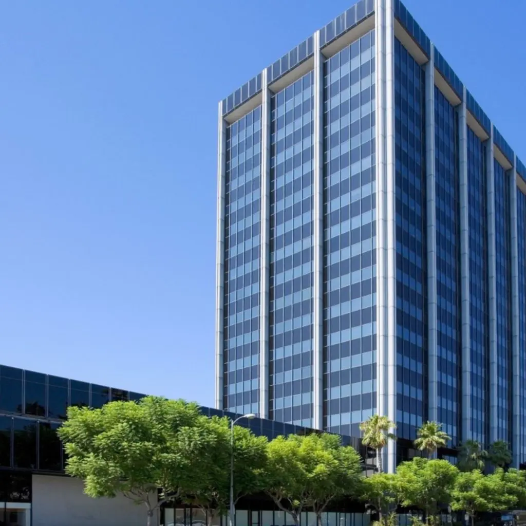 Barrister Suites Office Lease Tall modern office building with a glass facade reflecting blue sky, adjacent to a lower structure, with green trees and palm trees in the foreground.