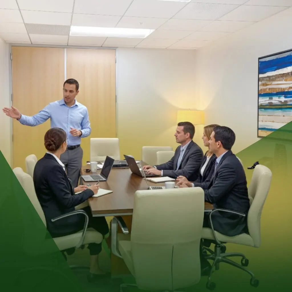 Barrister Suites Office Lease A man in a blue shirt stands and gestures while speaking to four colleagues seated around a conference table in a modern office meeting room with laptops and notepads.