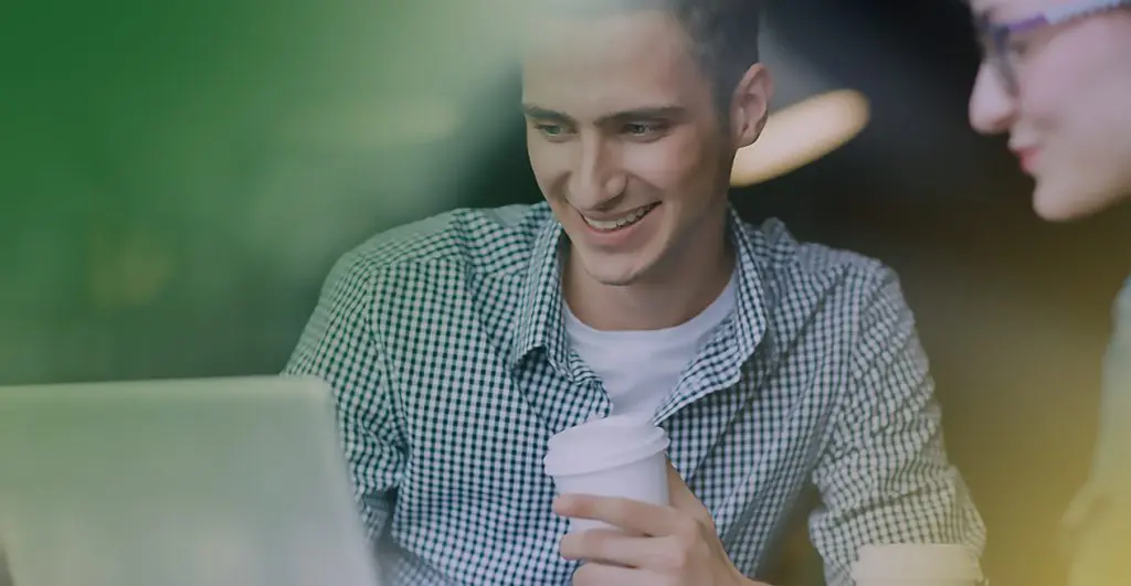 Barrister Suites Office Lease A young man smiling while looking at a laptop, holding a coffee cup. Another person sits nearby, slightly out of focus. The background is blurred, creating a warm and casual atmosphere.