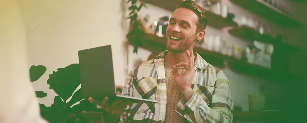 Barrister Suites Office Lease A smiling man in a plaid shirt holds a laptop and gestures with his hand while video chatting in a cozy, plant-filled room with shelves in the background.
