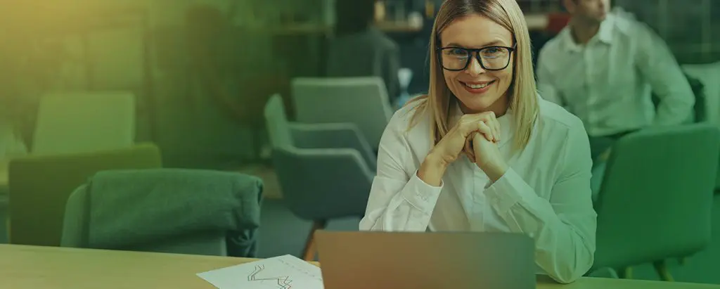 Barrister Suites Office Lease A woman with glasses and blonde hair, wearing a white shirt, sits at a desk with a laptop and papers, smiling at the camera in a modern office setting.