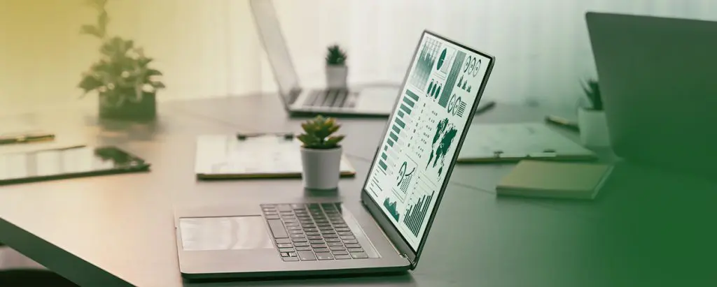 Barrister Suites Office Lease A laptop on a desk displays graphs and charts. The desk also has small potted plants, a clipboard, and another laptop in the background, with natural light coming through sheer curtains.